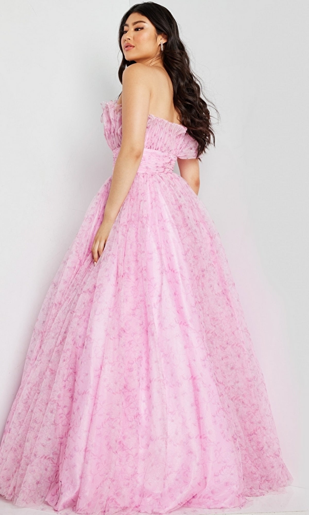 A woman wears the Jovani 26209 long prom dress, a strapless pink tulle A-line gown with a fitted bodice, full skirt, and subtle ruffle detailing. The floor-length design stands out elegantly against a plain light background.